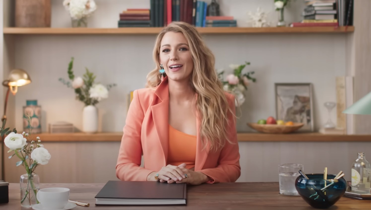 Blake Lively (actress) in a pink suit sitting at the desk with flowers, a cup, and a notebook