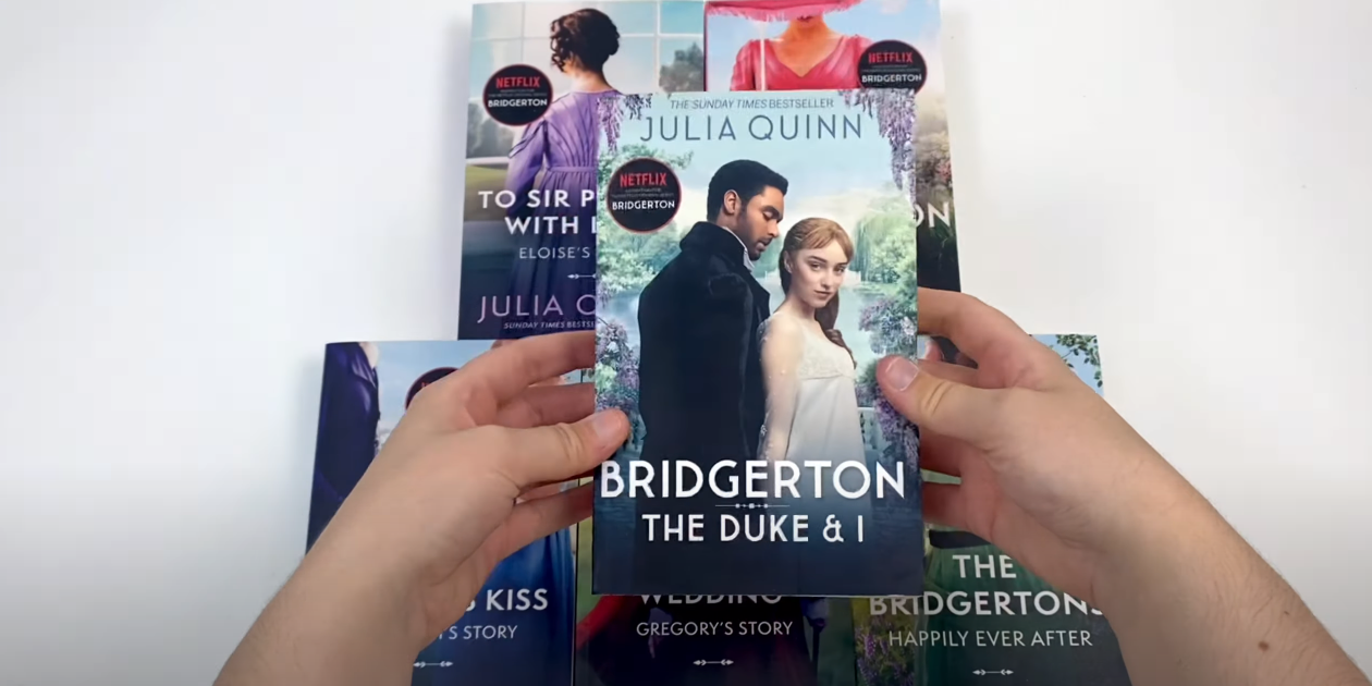 Hand Holding Bridgerton Book with Books on Table in Background